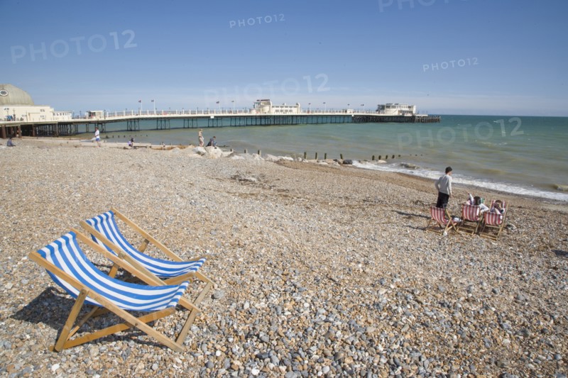 ENGLAND, West Sussex, Worthing, View across beach towards the pier with blue and white deckchairs in the foreground and sunbathers enjoying the sunshine nearby