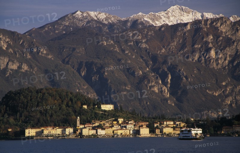 ITALY, Lombardy, Lake Como, Ballagio. View across Lake Como towards distant town of Ballagio situated at foot of tree covered hillside with mountain backdrop. Car ferry crossing lake in foreground.