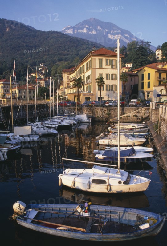 ITALY, Lombardy, Lake Como, Menaggio.  Harbour with moored boats and waterside buildings with tree covered mountain backdrop.