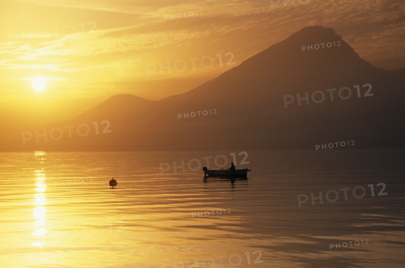 ITALY, Lake Garda, Fishing boat on lake at sunset in golden light with mountain peak in haze of sunshine behind.