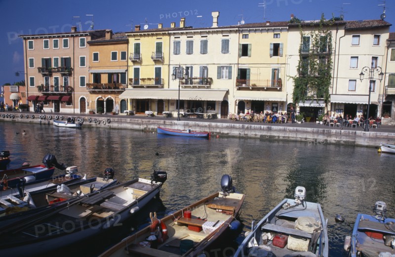 ITALY, Veneto, Lake Garda, Bardolino. Harbour scene with people at outside tables of waterside cafes and line of moored boats in foreground.  Pastel coloured building facades reflected in the water.