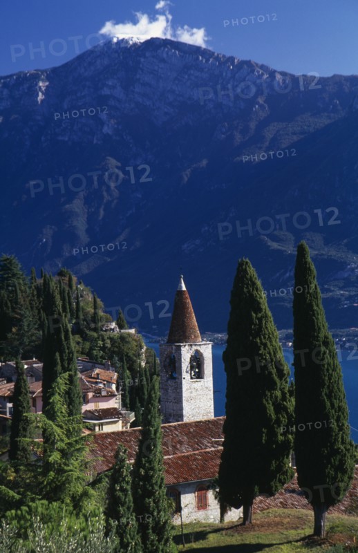 ITALY, Lombardy, Lake Garda, "Pieve.  View of tiled rooftops, church and bell tower of village overlooking Western shore of Lake Garda.  Cypress trees in foreground and mountain backdrop."
