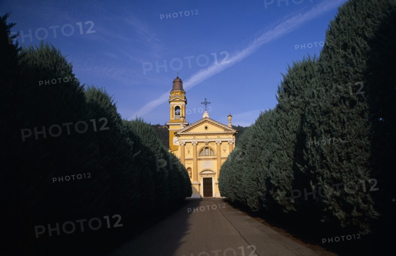 ITALY, Lombardy, Yellow and white exterior facade of small church with bell / clock tower at side with tree lined road leading to entrance.