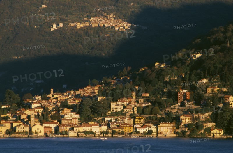 ITALY, Lombardy, Lake Como, Torno.  View across Lake Como towards town spread across hillside in golden sunlight.