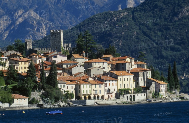 ITALY, Lombardy, Lake Como, Rezzonico.  View across lake to red tiled rooftops of village houses with castle tower and walls behind.