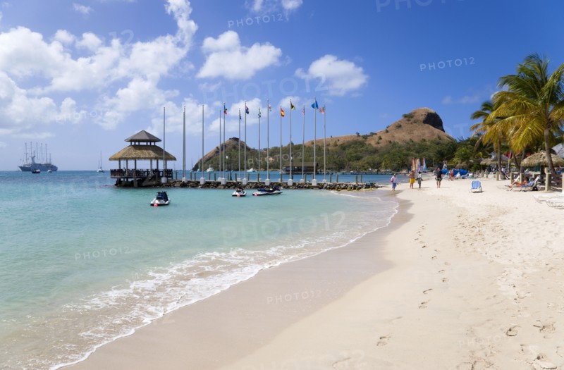 WEST INDIES, St Lucia, Gros Islet , Tourists on the beach at Sandals Grande St Lucian Spa and Beach Resort hotel beside a wooden jetty with Pigeon Island National Historic Park beyond