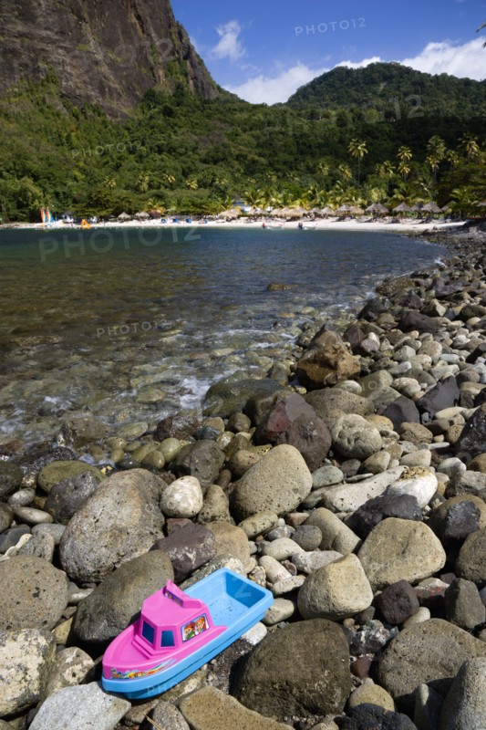 WEST INDIES, St Lucia, Soufriere , Val des Pitons The white sand beach at the Jalousie Plantation Resort Hotel with the volcanic plug of Petit Piton beyond and tourists on sunbeds beneath palapa sun shades with a childs plastic boat marooned on rocks in the foreground