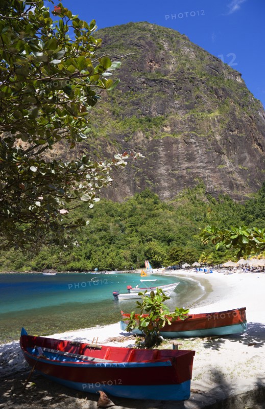 WEST INDIES, St Lucia, Soufriere , Val des Pitons The white sand beach at the Jalousie Plantation Resort Hotel with the volcanic plug of Petit Piton beyond and tourists on sunbeds beneath palapa sun shades with colourful fishing boats under a tree in the foreground