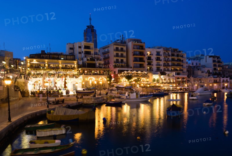MALTA, Saint Julians, Saint Julians Bay waterfront at dusk with illuminated restaurants in front of apartments and the Hilton Tower office complex with fishing boats in the harbour in the foreground