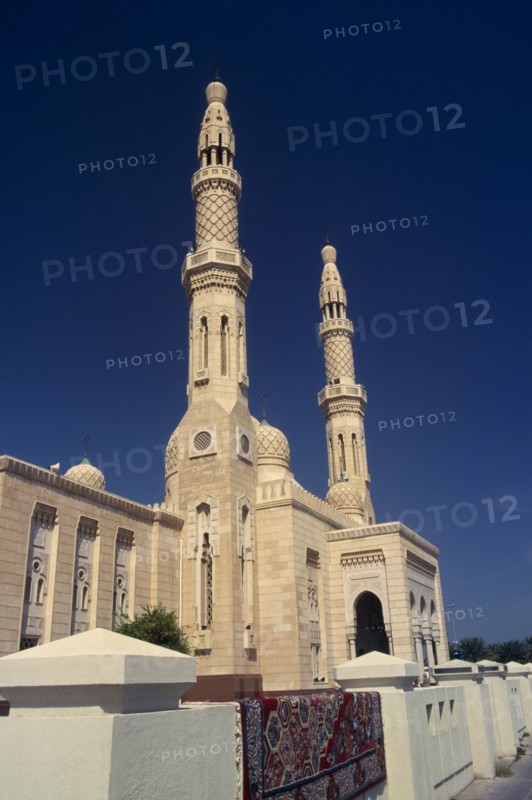 UAE, Dubai, Jumeirah Mosque.  Angled view of exterior with pair of minarets and domed rooftop decorated with hexagonal pattern.  Carpet lying over white wall in foreground.