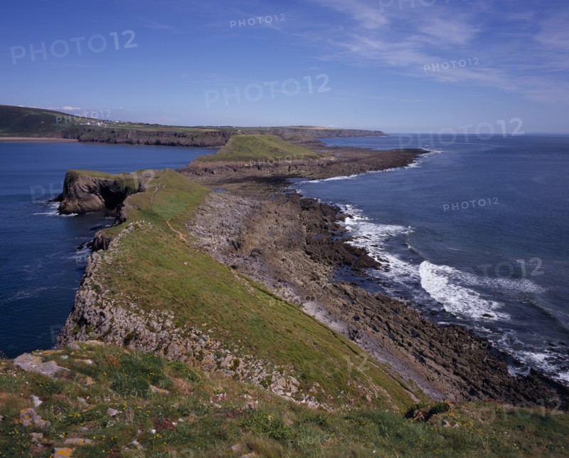 WALES, Swansea, Gower Peninsula, View east from the Worm’s Head towards the mainland.  Sloping ridge of cliff extending from mainland with waves breaking against side.