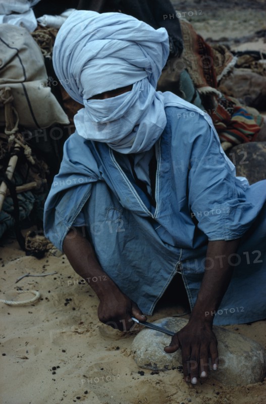 ALGERIA, Sahara Desert, Tuareg, "Tuareg man sharpening tool on rock, wearing blue taguelmoust covering face and head except for eyes. In Tuareg societies men are veiled rather than women.   "