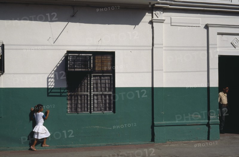 NICARAGUA, Leon, Street scene with little girl in white outside building painted dark green and white.  Metal window grill casting shadow above and man standing in doorway on right.
