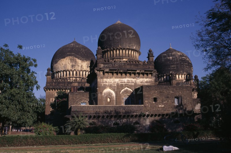 INDIA, Karnataka, Bijapur, Unidentified old building in Islamic architectural style with three domes set in gardens or green space with person asleep beside bicycle in foreground.