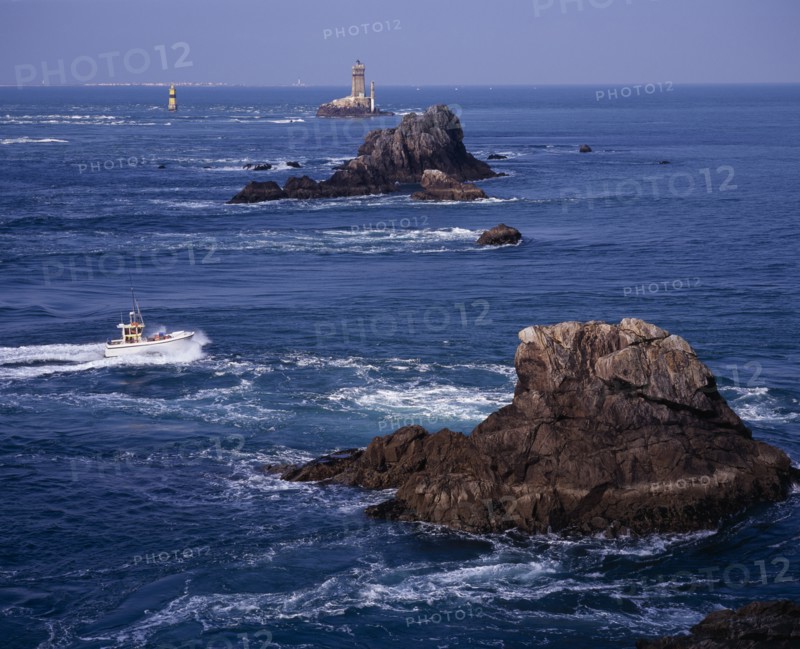 FRANCE, Brittany, Finistere, "Pointe du Raz.  Jagged outcrops of offshore rock protruding from sea with small fishing boat facing oncoming tide, light house and Ile de Sein skyline."