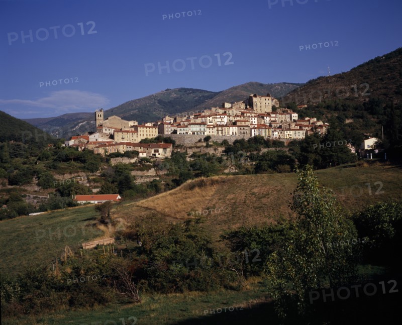 FRANCE, Languedoc-Roussillon, Pyrenees-Orientales, Mosset. View towards hillside village from the east with narrow houses with painted window shutters and red tiled rooftops. Backdrop of rocky hills scatteres with trees.