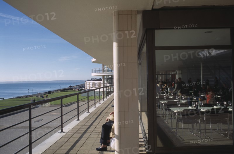 ENGLAND, East Sussex, Bexhill-on-Sea, De La Warr Pavilion. View along the sun terrace next to the restaurant with people sitting on benches behind a pillar looking out to the sea.