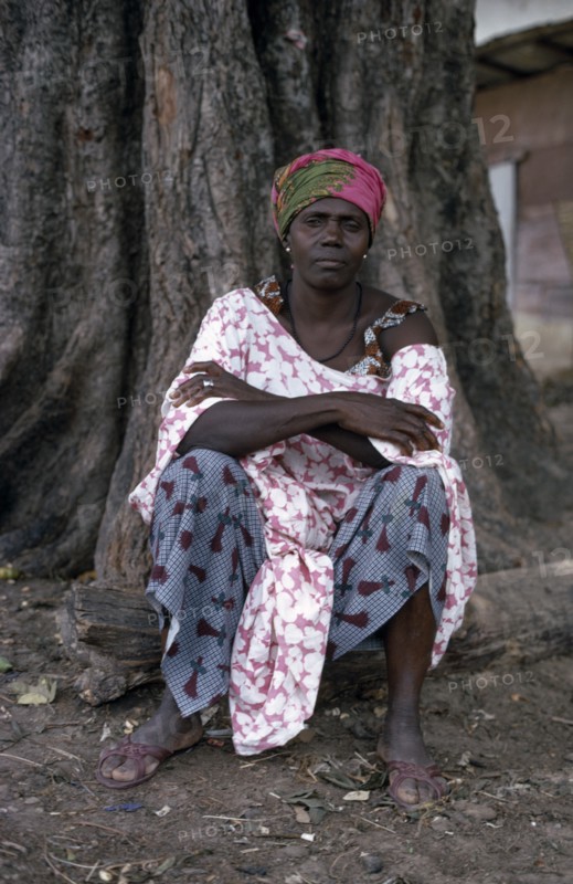SENEGAL, People, Portrait of woman seated at base of tree trunk wearing fabric of different printed patterns.