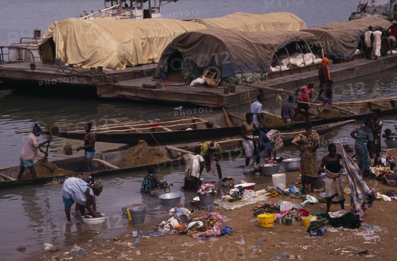 SENEGAL, Niger River, Women washing clothes in river from bank with barges and wooden canoes transporting soil and other goods behind.