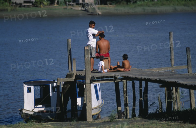 20088598 BRAZIL Para Aflua Amazon Estuary.  Man having a haircut while waiting for the ferry on wooden jetty watched by two children.