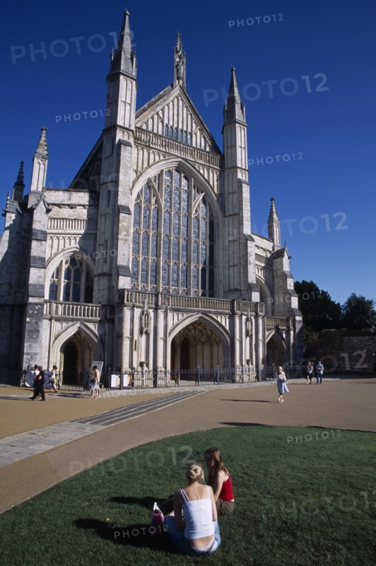 ENGLAND, Hampshire, Winchester, Winchester Cathedral exterior seen from the gardens with two girls sat on the grass in the foreground.