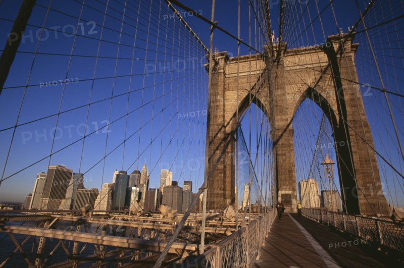 USA, New York, New York City, "Brooklyn Bridge.  View across bridge, pedestrians and cyclist to Manhattan skyline part framed by stone tower and intersected by steel wires and suspension cables. Spans the East River.  Construction began in 1870 and the bridge opened for use in 1883."