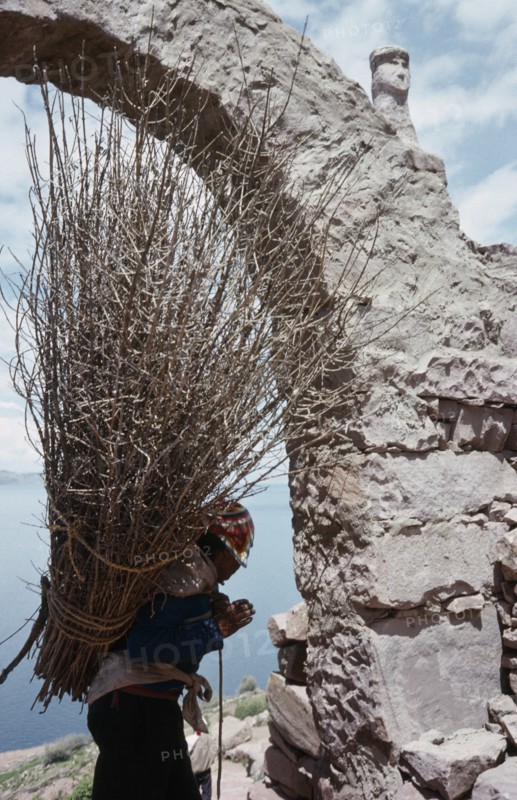 PERU, Puno, Lake Titicaca, Taquile Island.  Local man carrying bundle of twigs on his back framed in partly seen stone archway.