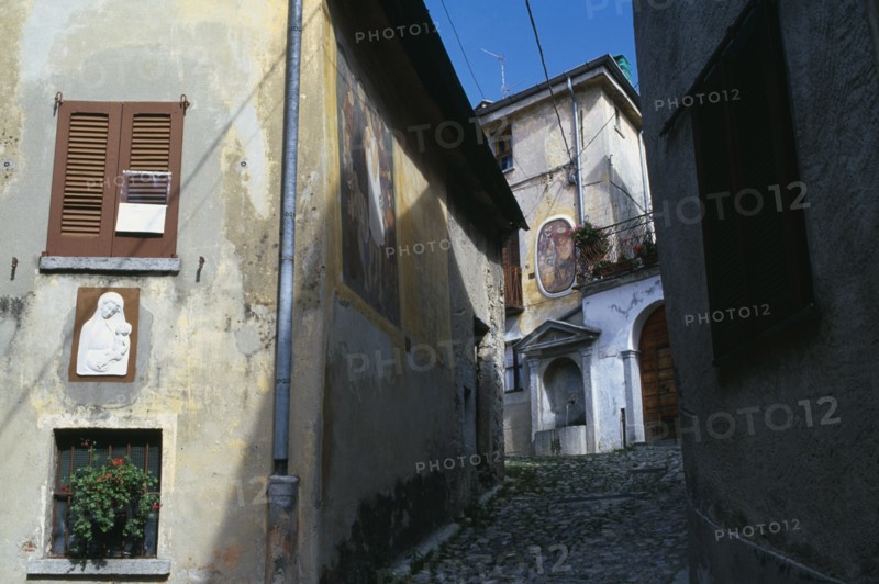 ITALY, Lombardy, Arcumeggia, Narrow cobbled street with fresco decoration and plaster relief of the Virgin and Child on the exterior walls of the buildings and drinking fountain at top.