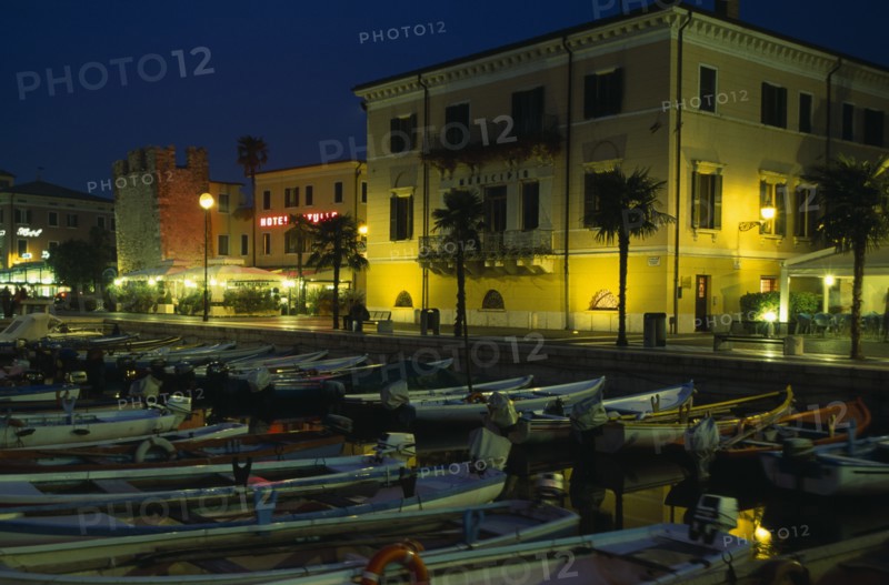 ITALY, Veneto, Lake Garda, Bardolino.  Harbour and moored boats at night with waterside bars and hotels and illuminated street lights and signs.