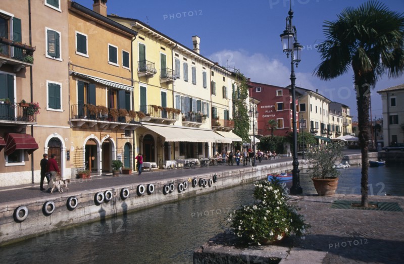 ITALY, Veneto, Lake Garda, Bardolino.  Harbour and line of waterside bars and cafes painted in pastel colours and with awning pulled out over outside tables.  Moored boats in foreground.
