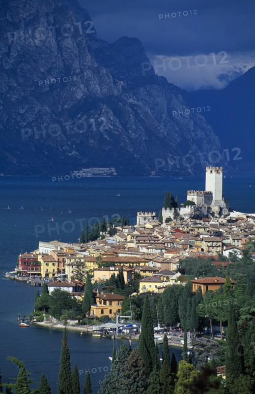 ITALY, Veneto, Lake Garda , Malcesine.  Tiled rooftops and pastel coloured buildings of town and castle on shore of lake with mountains beyond.