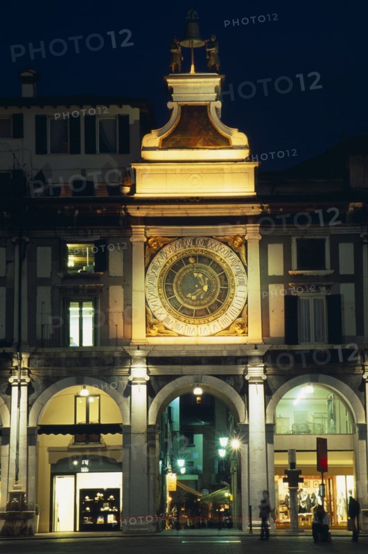 ITALY, Lombardy, Brescia, "Astronomical clock in Piazza della Loggia illuminated at night with brightly lit shop windows framed by archways of colonnade below.  People, street lamps and light trail."