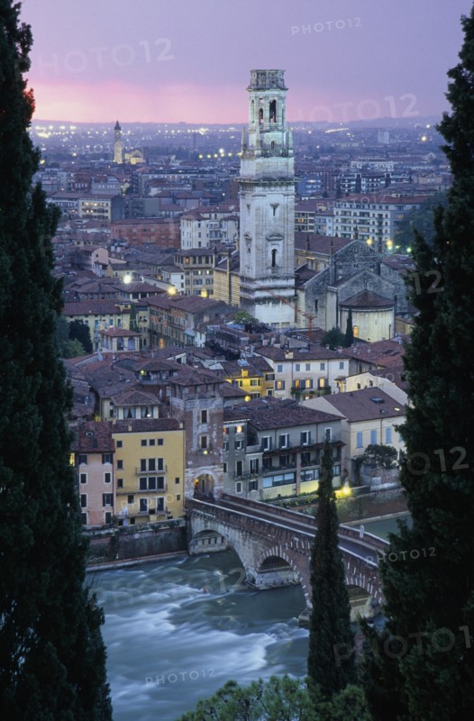 ITALY, Veneto, Verona, "Cityscape at sunset showing tiled rooftops, Duomo bell tower and bridge over the Adige River part framed by tall coniferous trees.  Pale pink/purple sky and city lights."