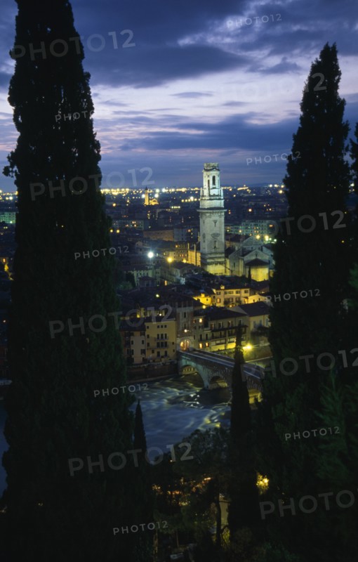 ITALY, Veneto, Verona, "Cityscape illuminated at night showing tiled rooftops, Duomo bell tower and bridge over the Adige River part framed by tall coniferous trees.  Windswept evening sky."
