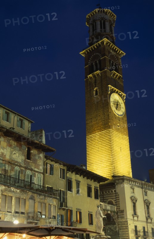 ITALY, Veneto, Verona, "Torre dei Lamberti clock tower in the Piazza Erbe illuminated at night part seen behind faded building facades, statue and lights and umbrellas of cafe bar."