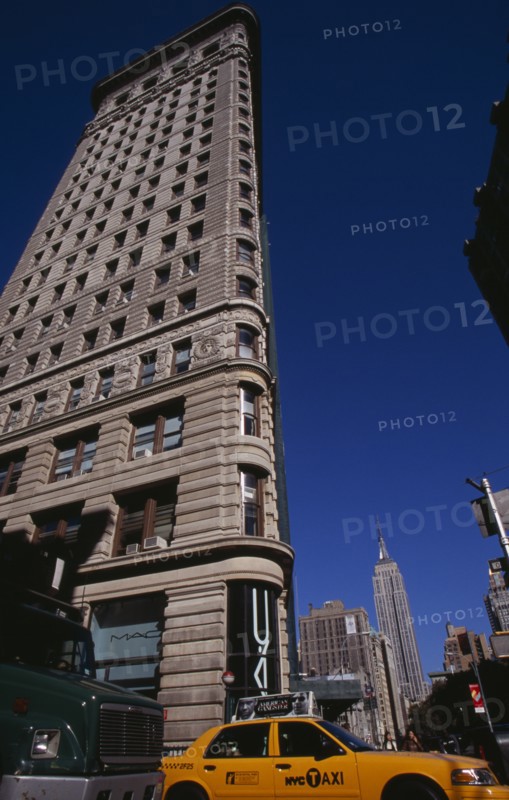 USA, New York, New York City, "Angled view of the Flatiron Building from 23rd Street.  Traffic including yellow taxi cab below and Empire State Building beyond. Steel framed, Beaux-Arts skyscraper designed by architect Daniel Burnham in 1902 to a triangular plan. "