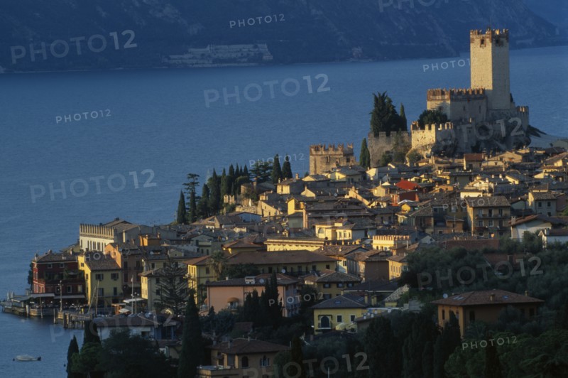 ITALY, Veneto, Lake Garda, "Malcesine.  View across town rooftops towards Castello Scaligero in warm, golden light with lake beyond."