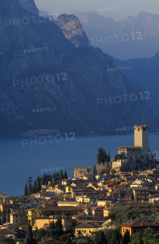 ITALY, Veneto, Lake Garda, "Malcesine.  View across town rooftops towards Castello Scaligero in warm, golden light with lake and mountains beyond."