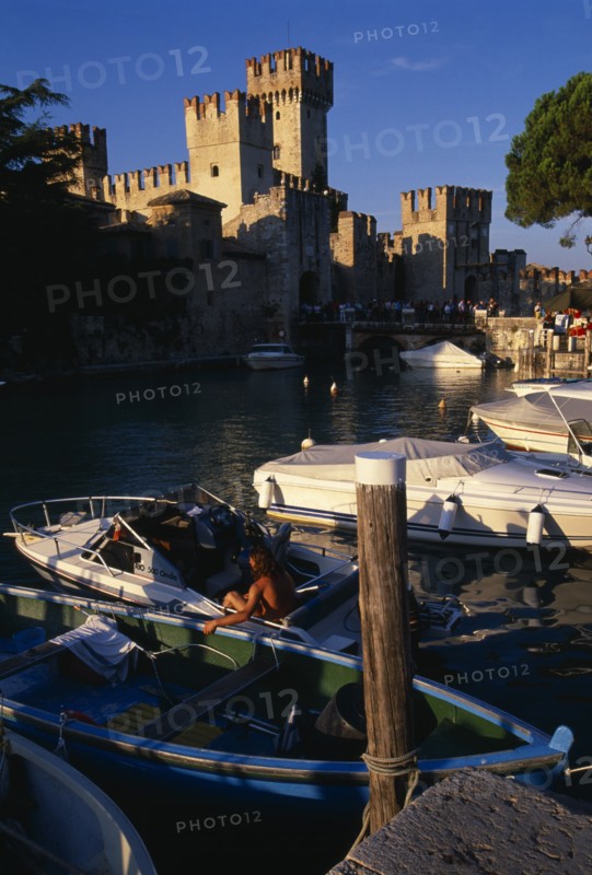 ITALY, Lombardy, Lake Garda , Sirmione.  Rocca Scaligera medieval castle with crowds of visitors crossing bridge to entrance.  Moored boats in foreground.