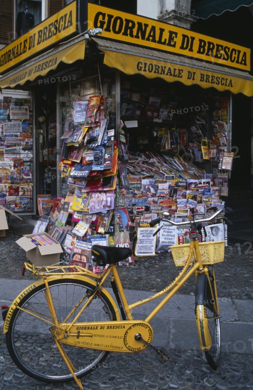 ITALY, Lombardy, Brescia, Piazza della Loggia.  Newspaper stand with yellow painted bicycle advertising name.