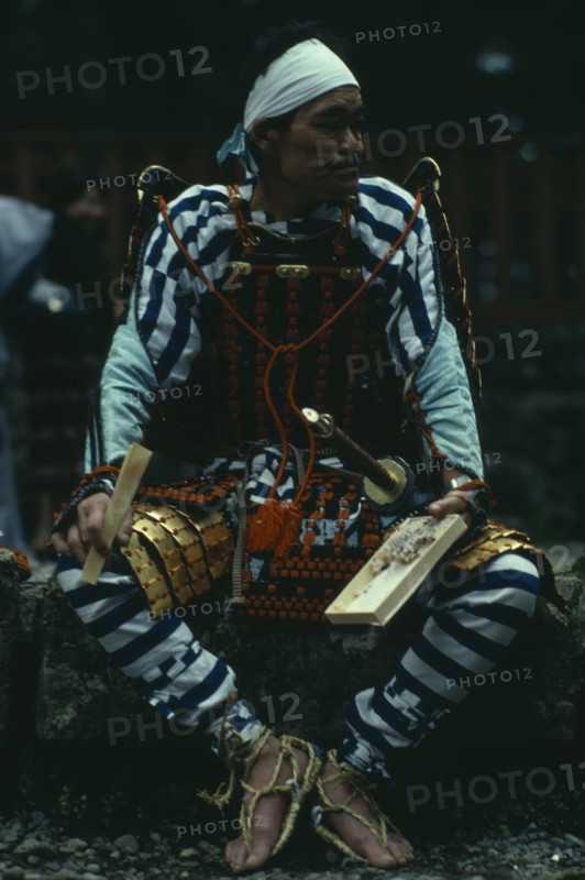 JAPAN, People, Samurai, "Portrait of Samurai wearing armour constructed of plates of metal or bamboo held together with coloured lacing, rope sandals and white cloth head band with sword at side.  Seated, holding bento lunchbox and chopsticks.  "