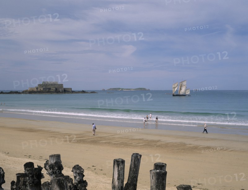 FRANCE, Brittany, St Malo, Le Sillon Beach and Fort National. People walking along golden sand with a sail boat on seen on water