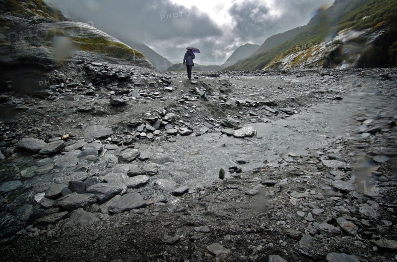NEW ZEALAND, SOUTH ISLAND, WEST COAST, "MOUNT COOK NATIONAL PARK, A WALKER CROSSES A STREAM INFRONT OF THE TERMINAL FACE OF THE FRANZ JOSEF GLACIER. Julius von Haast, geologist and explorer, named Franz Josef Glacier in 1863, after the Emperor of the Austro-Hungarian Empire. Approximately 7000 years old, and a remnant of a much older and larger glacier which originally swept right to the sea, Franz Josef Glacier extends 12 kilometres from its three feeder glaciers in the high snow fields of the southern Alps. "