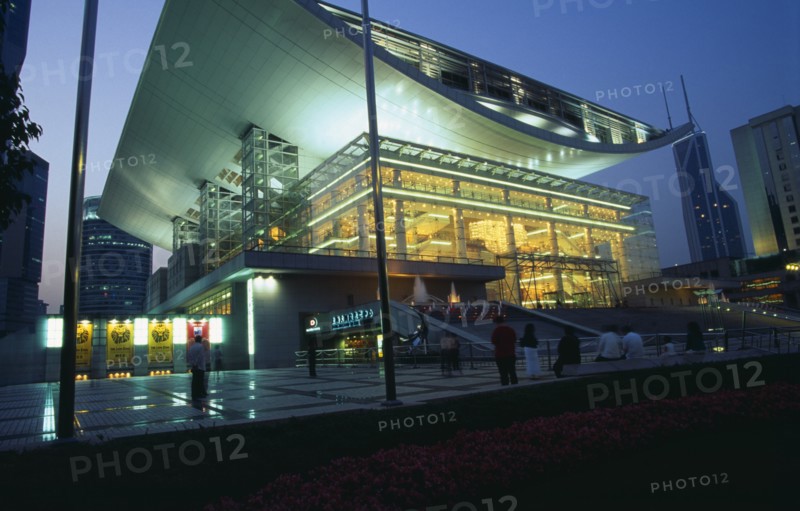 CHINA, Shanghai, "Opera House, Renmin Square.  Glass fronted exterior facade and upwardly curving roof illuminated at night with people standing or sitting in courtyard outside."