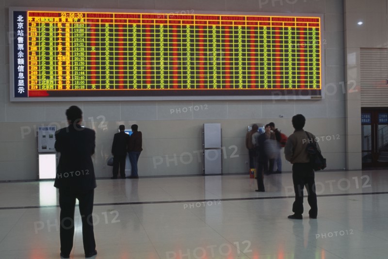 CHINA, Beijing, Interior of railway terminal with electronic information board with customers looking at smaller screens beside ticket machines below.