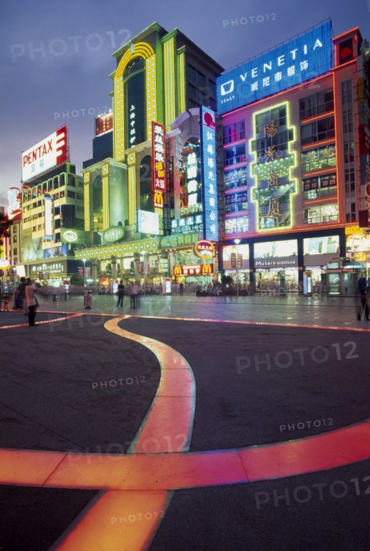 CHINA, Shanghai, Nanjing Lu.  Busy street scene at night with illuminated neon signs and advertising and ribbon of pink / orange lit pavement across square in the foreground.