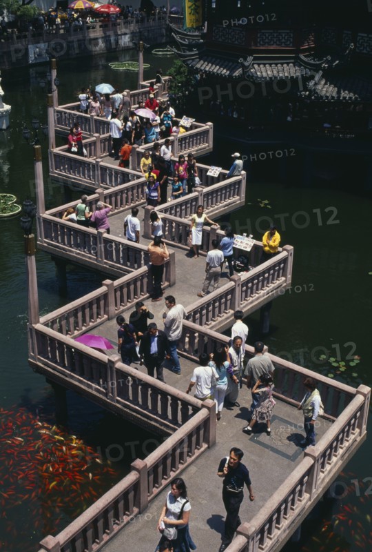 CHINA, Shanghai, "Yu Gardens.  View over zig-zag foot-bridge over pond with mass of silver and orange Koi carp. Vendors and crowds smoking, sightseeing and posing for / taking photograhs"