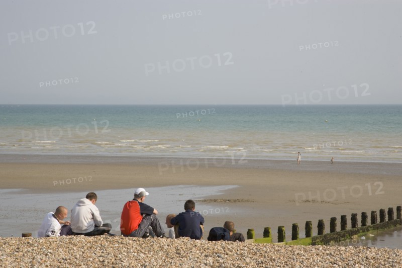 ENGLAND, West Sussex, Bognor Regis, A group of teenage boys sitting on shingle and sand beach looking out towards the sea