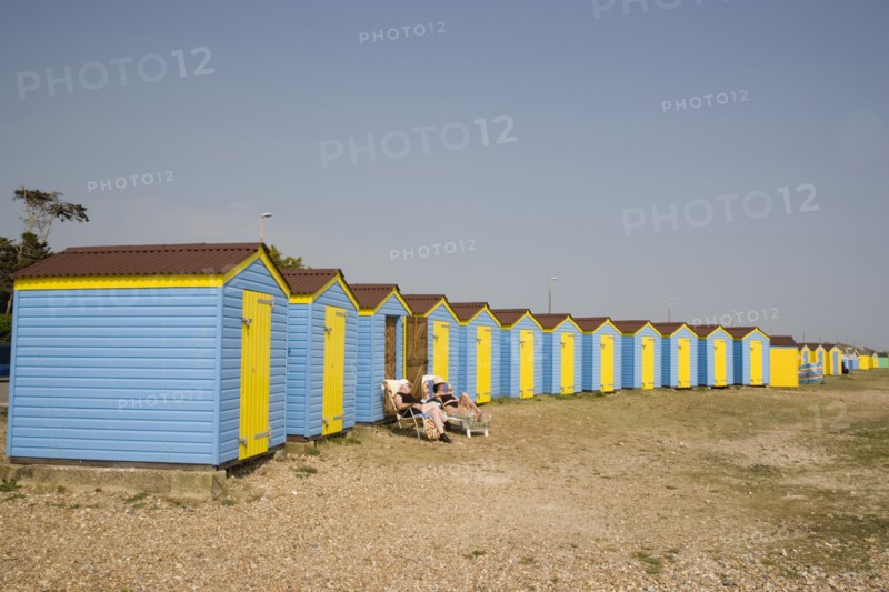 ENGLAND, West Sussex, Littlehampton, A crescent of blue and yellow beach huts with a couple sunbathing on loungers