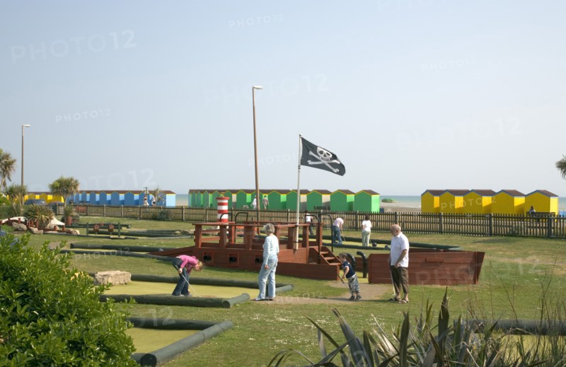 ENGLAND, West Sussex, Littlehampton, Families enjoying pitch and putt mini golf games in Norfolk Gardens next to seafront. A skull and cross bones flag flying. Colourful beach huts behind.